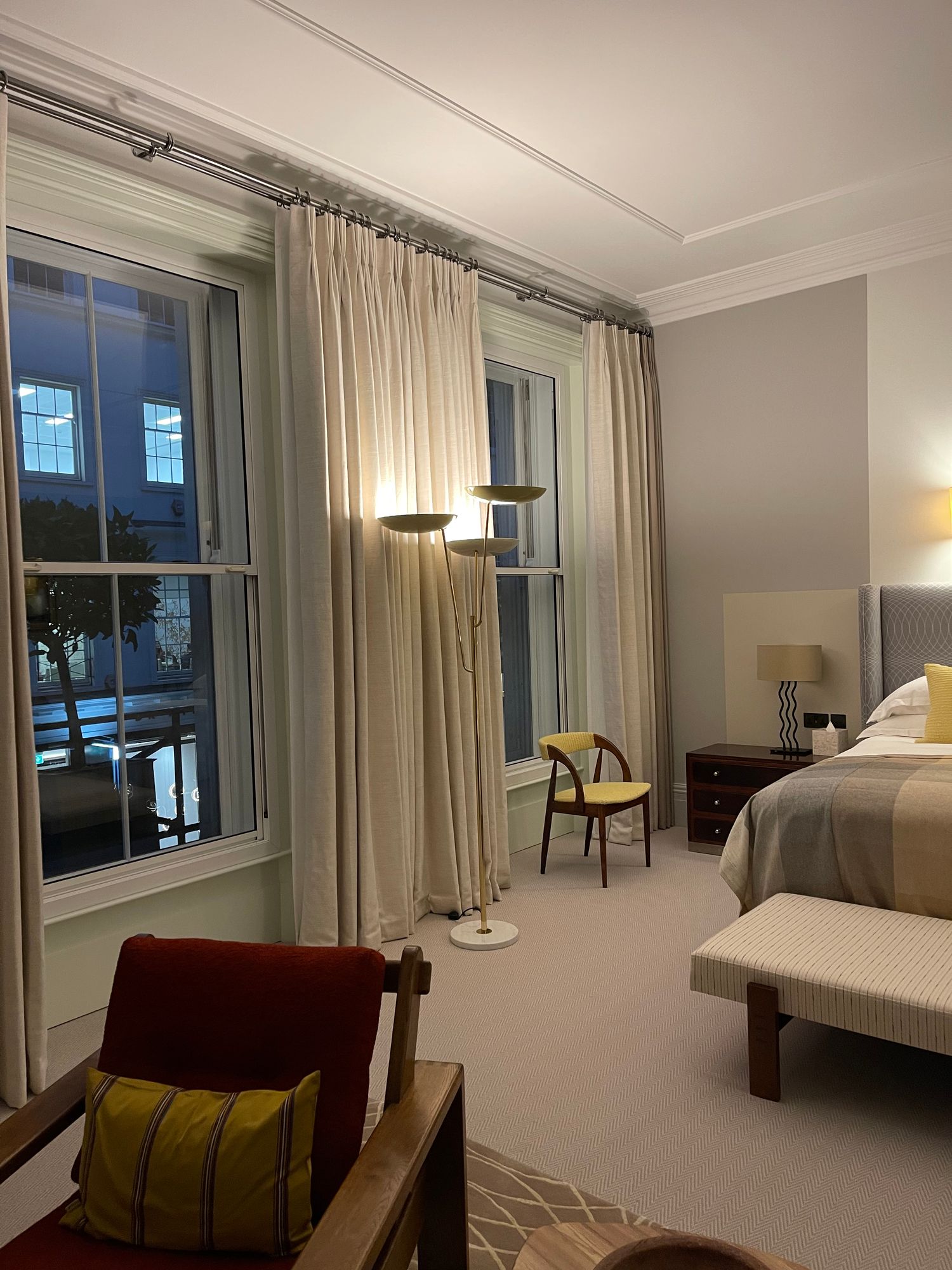 Guest bedroom at Brown's Hotel, Mayfair, with tall sash windows, full-length linen curtains, a red armchair and an upholstered bench at the foot of the bed.